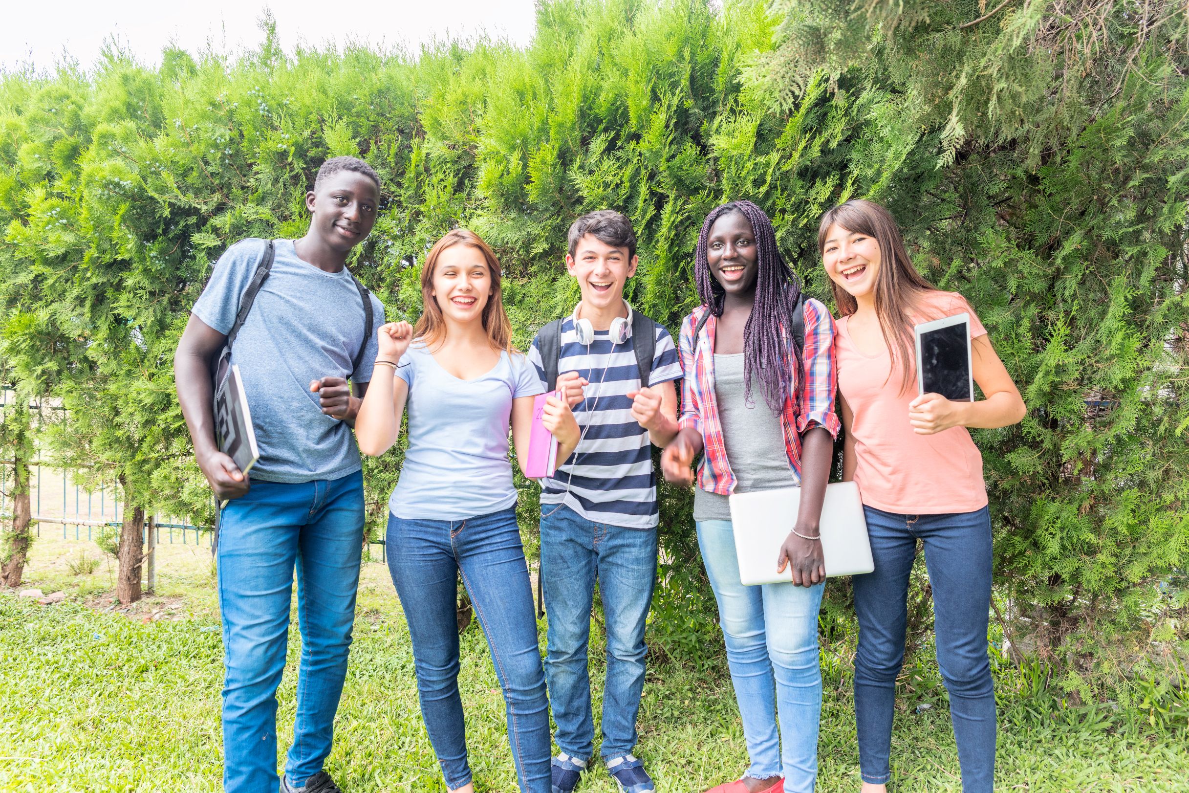 Five teenagers standing in park and smiling at the camera with notebooks and laptops.